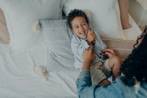 Sweet excited mixed race boy lying on bed and laughing while playing with his loving mom for article about mindful parenting for ADHD